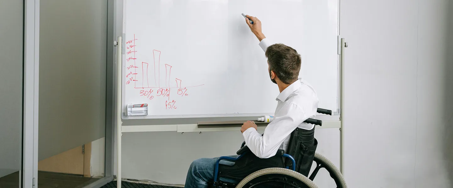 Ein im Rollstuhl sitzender Mann schreibt in einem Büro etwas auf ein Flipchart, auf dem sich bereits eine Grafik mit Zahlen befindet.