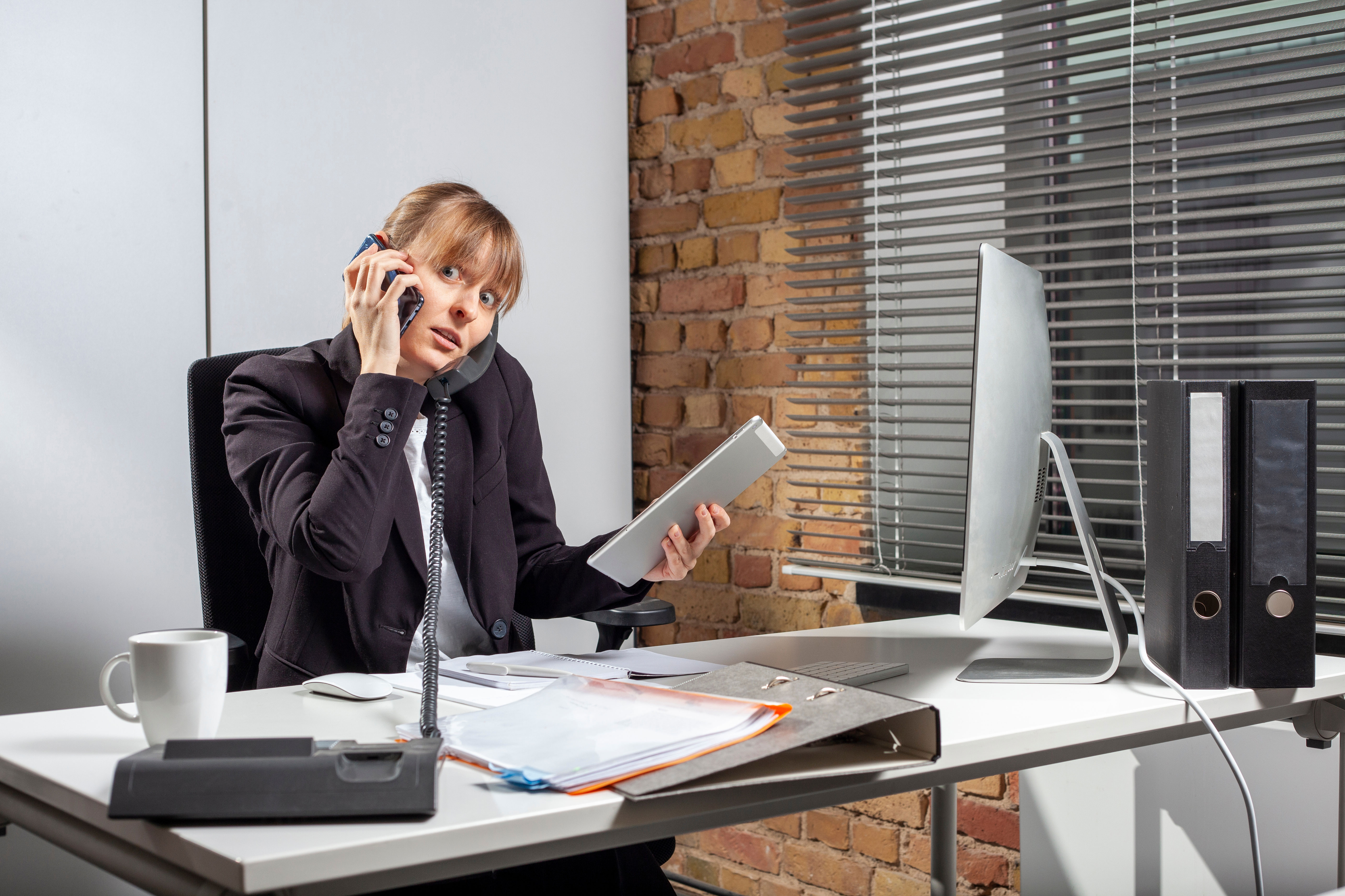 Gestresste Frau telefoniert auf Smartphone und Festnetztelefon während sie auf Tablet und Computer Eingaben tätigt. 