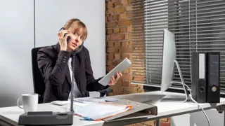 Gestresste Frau telefoniert auf Smartphone und Festnetztelefon während sie auf Tablet und Computer Eingaben tätigt. 