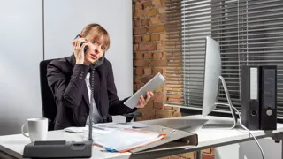 Gestresste Frau telefoniert auf Smartphone und Festnetztelefon während sie auf Tablet und Computer Eingaben tätigt.