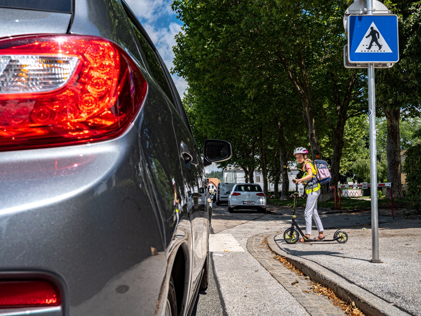 Blick vorbei an einem Pkw, der sich einem Fußgängerübergang nähert. Am Straßenrand steht ein Schulkind auf einem Tretroller und blickt in Richtung Pkw.