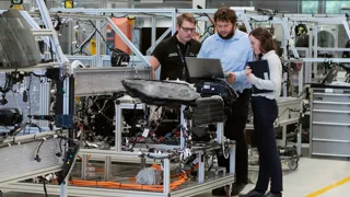 Zwei junge Männer und eine junge Frau schauen in einer Produktionshalle gemeinsam auf einen Laptop. Zwei junge Männer und eine junge Frau schauen in einer Produktionshalle gemeinsam auf einen Laptop.