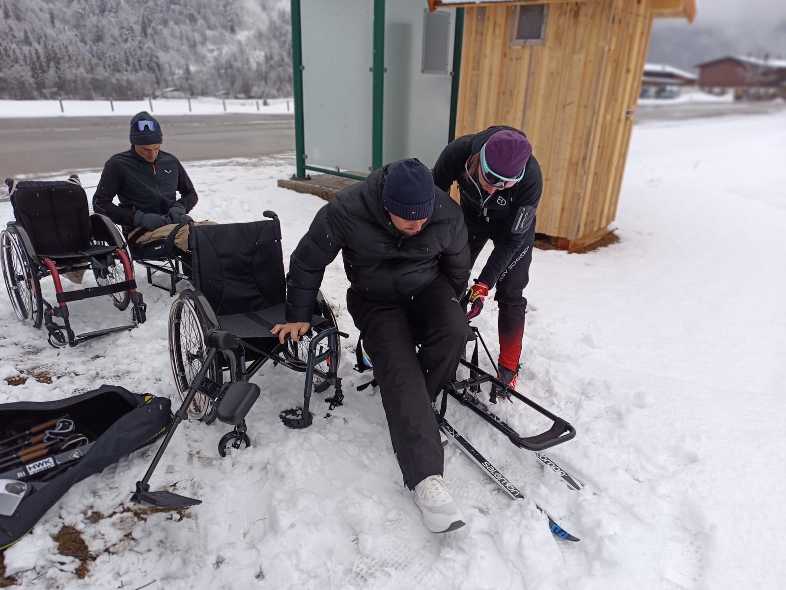 Die Trainerin hilft einem Teilnehmenden beim Umstieg aus dem Rollstuhl in den Langlaufschlitten 