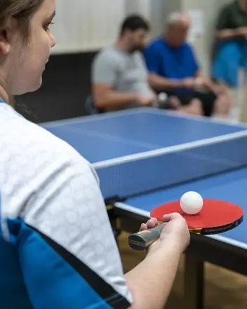 Eine Sportlerin mit Tischtennisschläger und Ball, im Hintergrund weitere Sportler