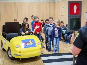 Mehreren jungen Schulkindern wird von einer Lehrerin, die in einem gelben Übungsauto sitzt, das richtige Verhalten an einer Ampel mit Zebrastreifen erklärt.
