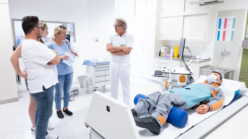 Ein Mitarbeiter des RH Häring spricht mit drei Besucher:innen in einem Behandlungsraum, auf der Liege liegt ein Patient, bei dem es sich jedoch zu Demonstrationszwecken um eine Puppe handelt