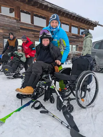 Teilnehmer:in mit Trainer vor der Skihütte, im Hintergrund weitere Teilnehmende mit Trainern