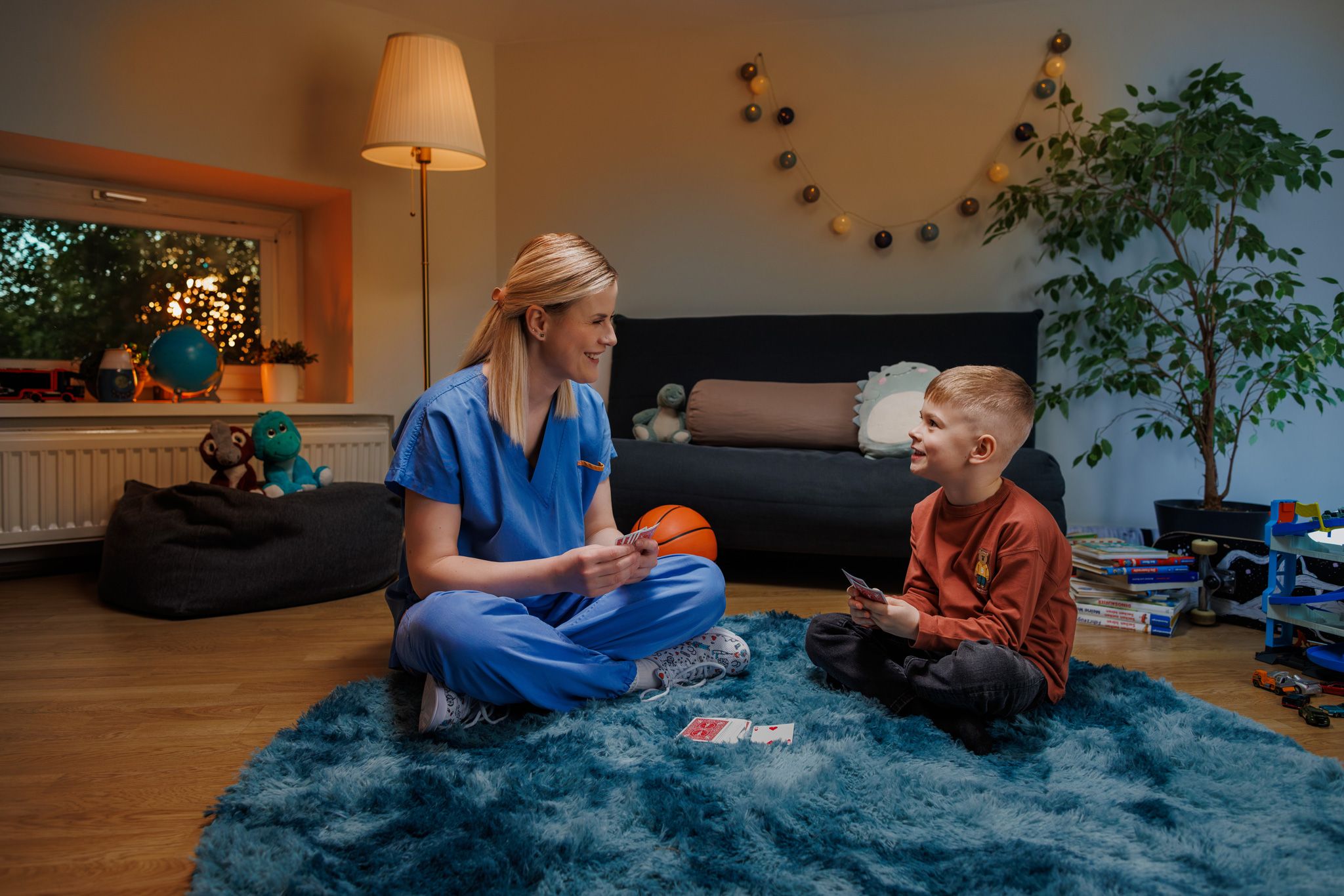 Eine Mutter sitzt mit hellblauer Krankenhaus-Dienstkleidung bekleidet mit ihrem Sohn in einem Kinderzimmer, sie spielen ein Kartenspiel.  