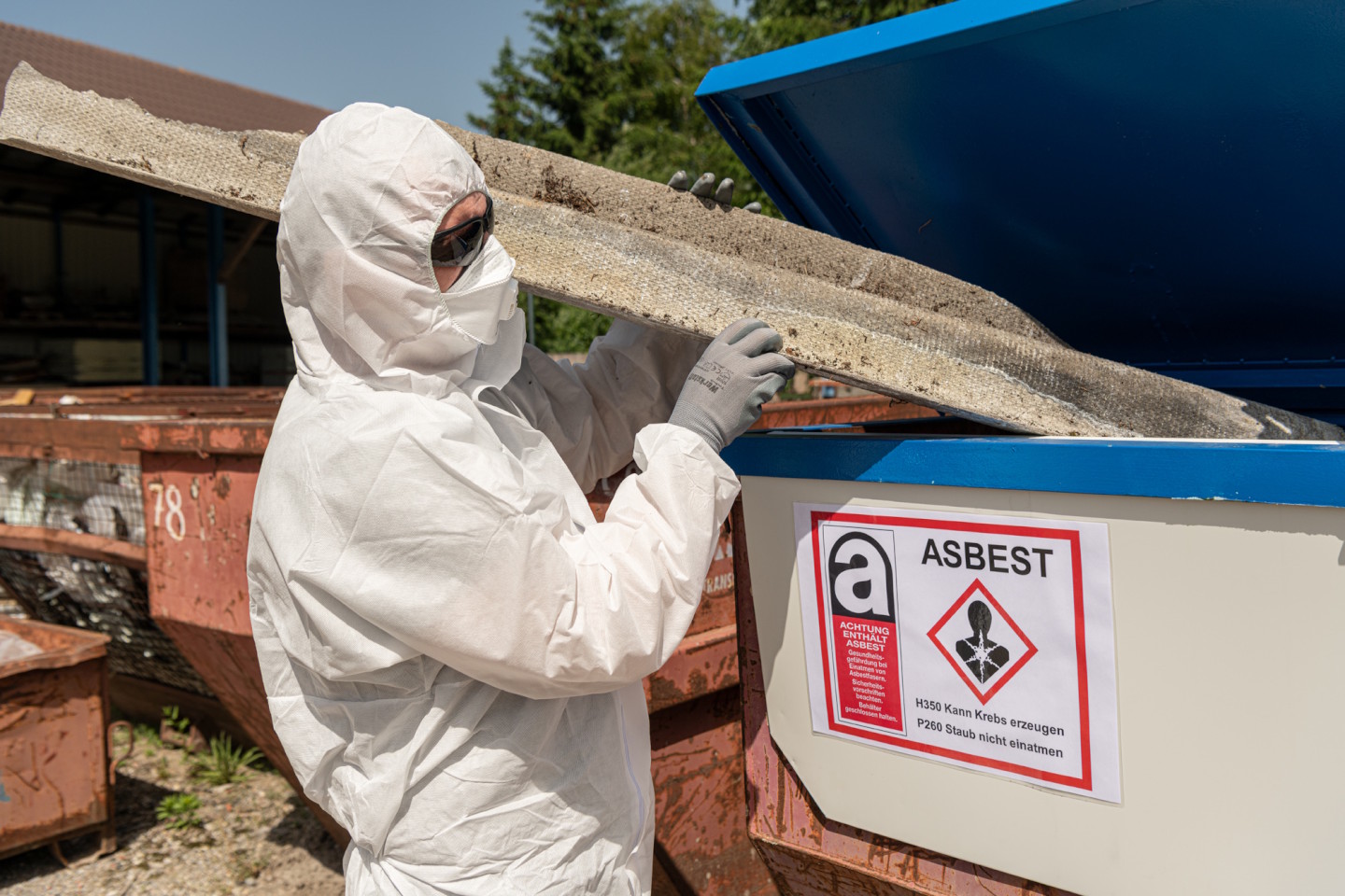 Mann in Schutzkleidung beim Beladen eines mit der Aufschrift Asbest gekennzeichneten Behälters 