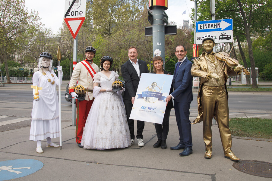 Sieben Personen stehen vor einer Straßenkreuzung und präsentieren die Aktion "Kluge Köpfe tragen Helm" von AUVA und KFV