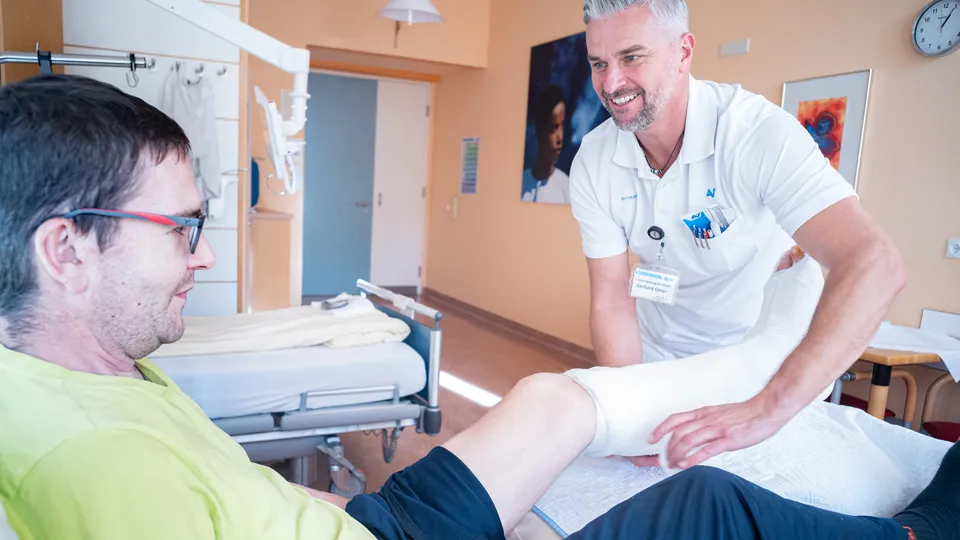 Ein Arzt behandelt in einem Krankenhauszimmer einen im Bett liegenden Patienten, der am linken Unterschenkel und Fuß einen Verband trägt.