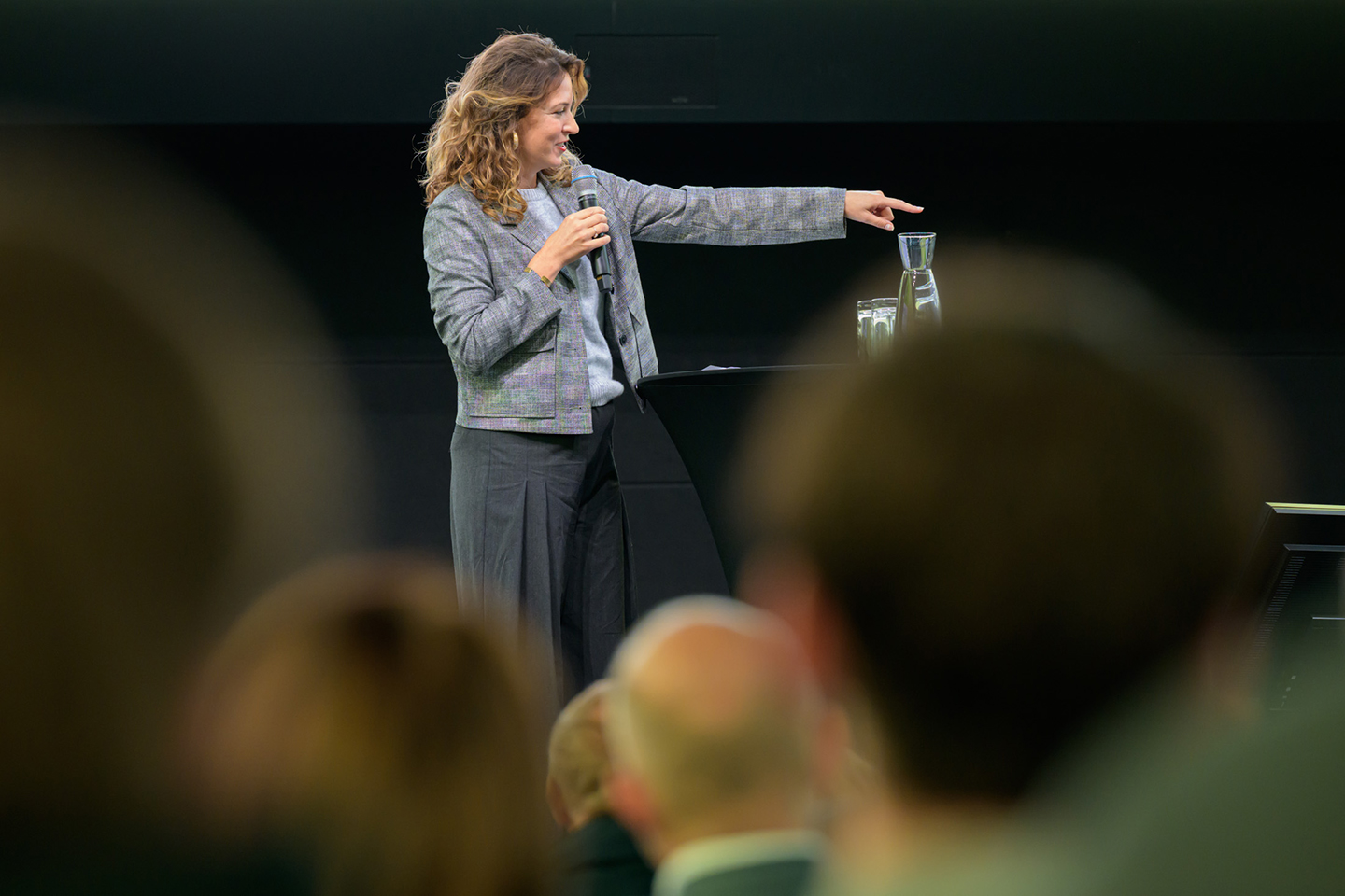 Ein Frau auf der Bühne deutet mit ausgestrecktem linken Arm auf etwas. In der rechten Hand hält sie ein Mikrofon
