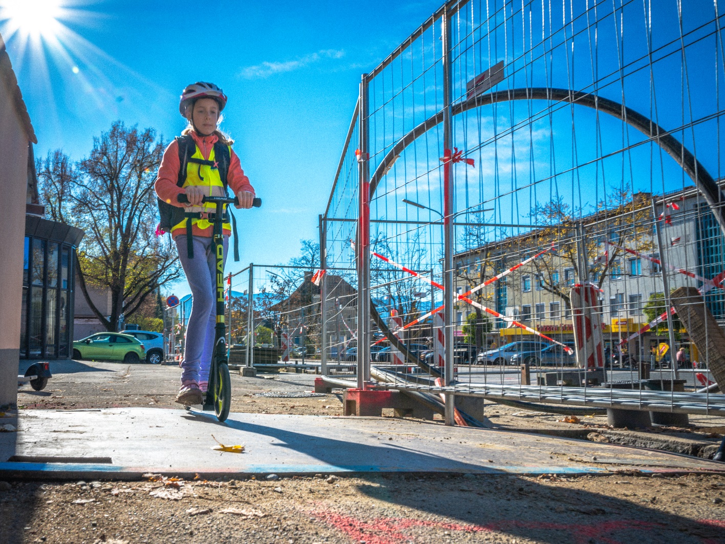 Ein Schulmädchen auf einem Tretroller fährt über eine Platte. Daneben sieht man die Absperrung einer Baustelle.