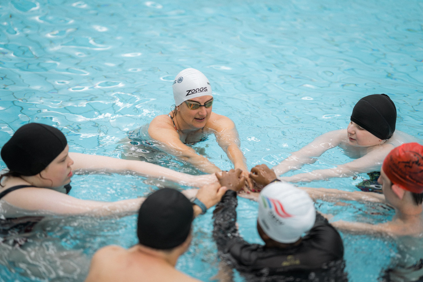 Sechs Teilnehmer:innen bilden im Schwimmbecken einen Kreis und legen ihre Hände aufeinander