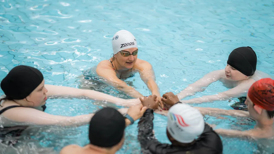 Sechs Teilnehmer:innen bilden im Schwimmbecken einen Kreis und legen ihre Hände aufeinander