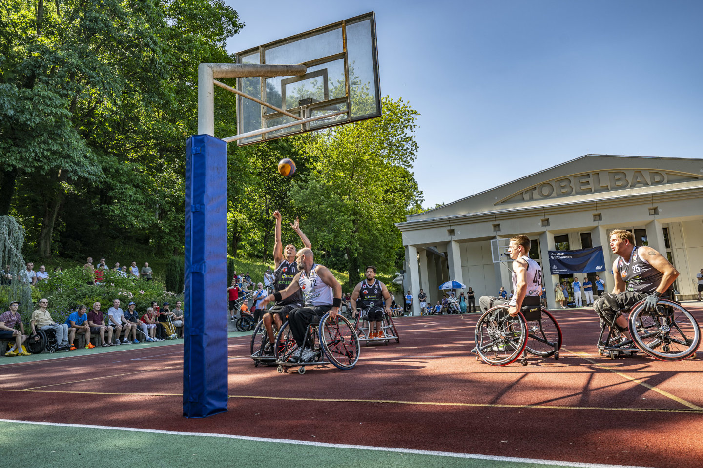 Mehrere Spieler mitten im Rollstuhlbasketballspiel, im Hintergrund Zuschauer 