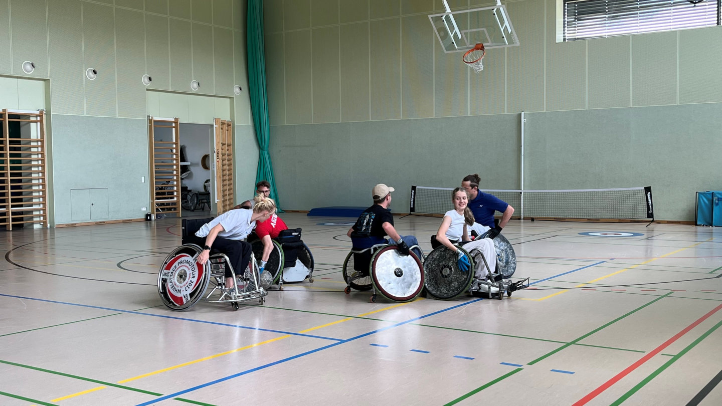 Fünf Rollstuhl-Rugby-Spieler:innen befinden sich in einem Turnsaal mitten im Spiel