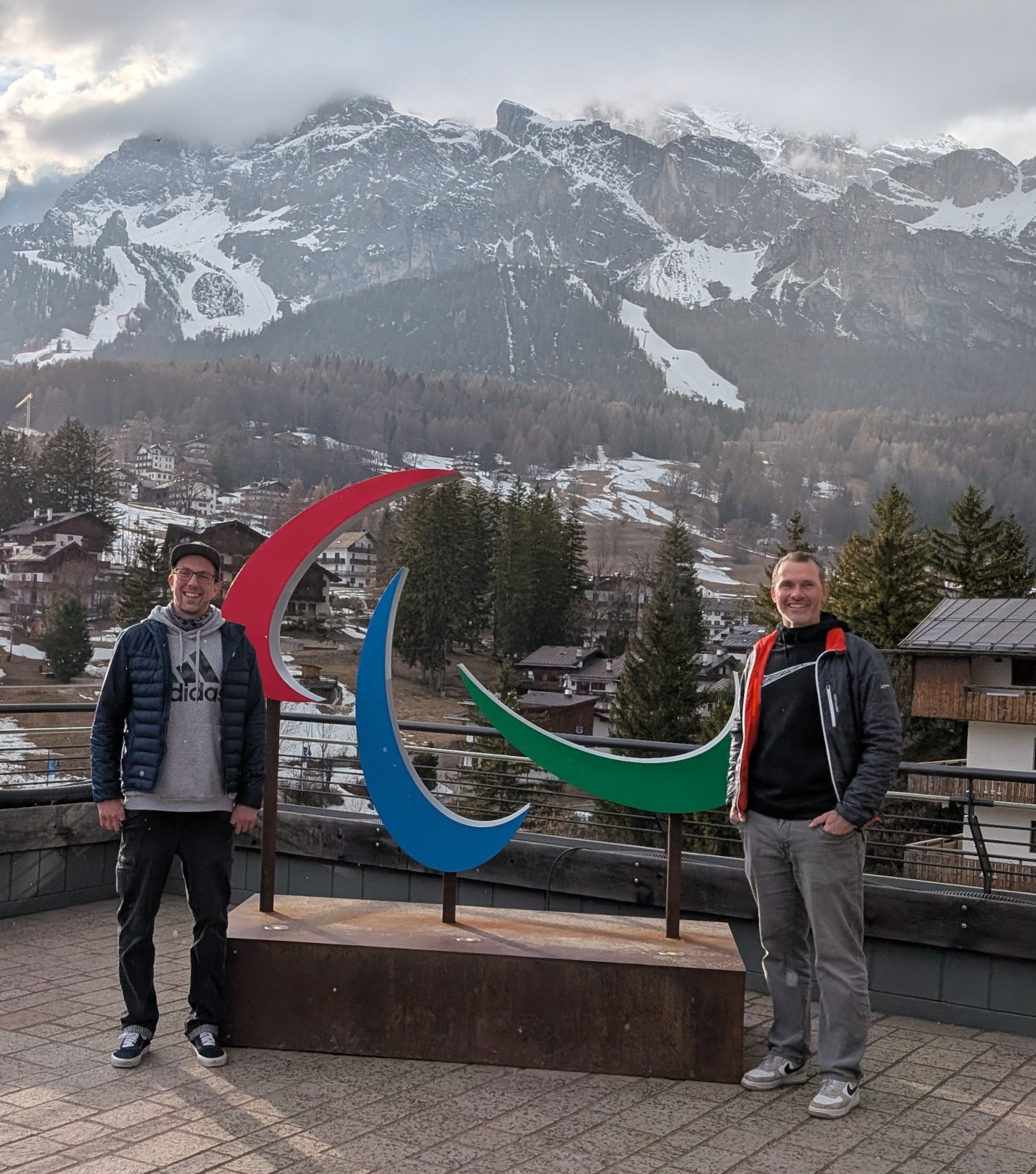 Zwei Mitarbeiter des Parasportzentrums Häring posieren neben einem großen Logo Paralympics-Logo, im Hintergrund eine winterliche Berglandschaft. 
