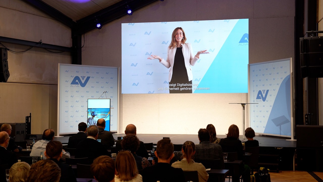 Blick aus dem Publikum auf die Bühne. Mittig ist auf einer großen Leinwand Caroline Krammer zu sehen.