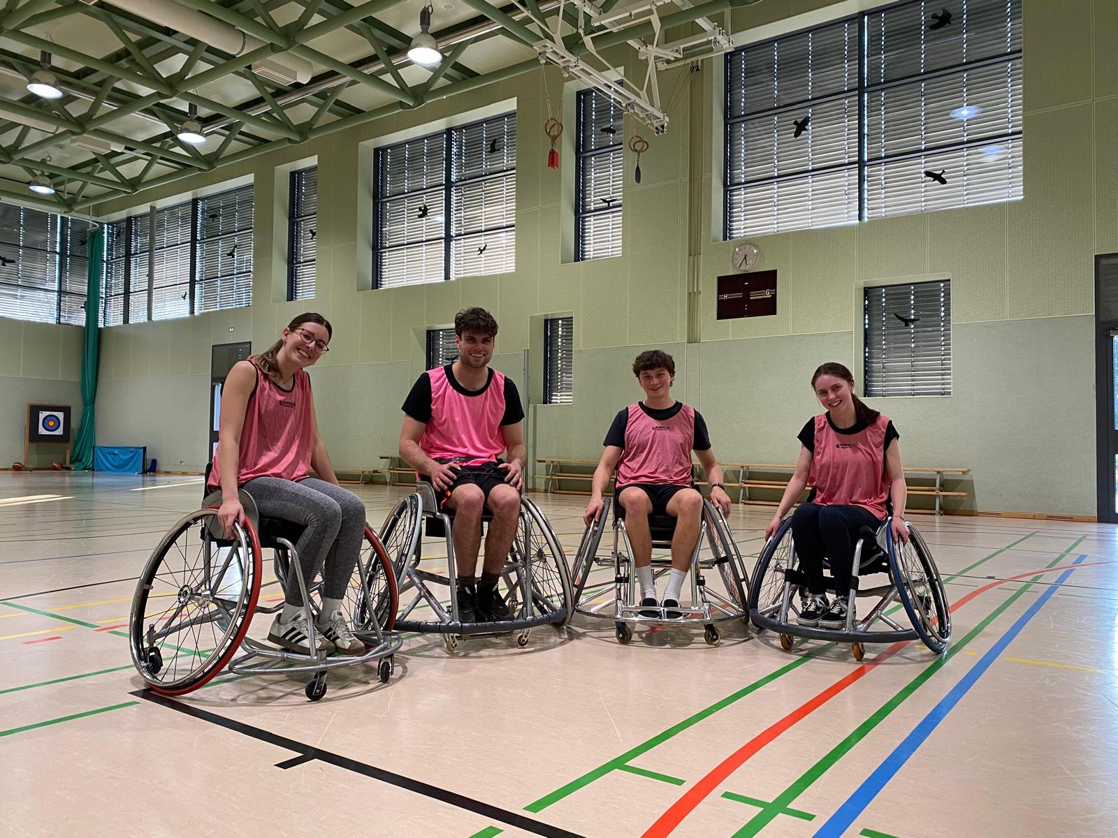 Vier Rollstuhlbasketballspieler:innen lächeln in einer Turnhalle in die Kamera
