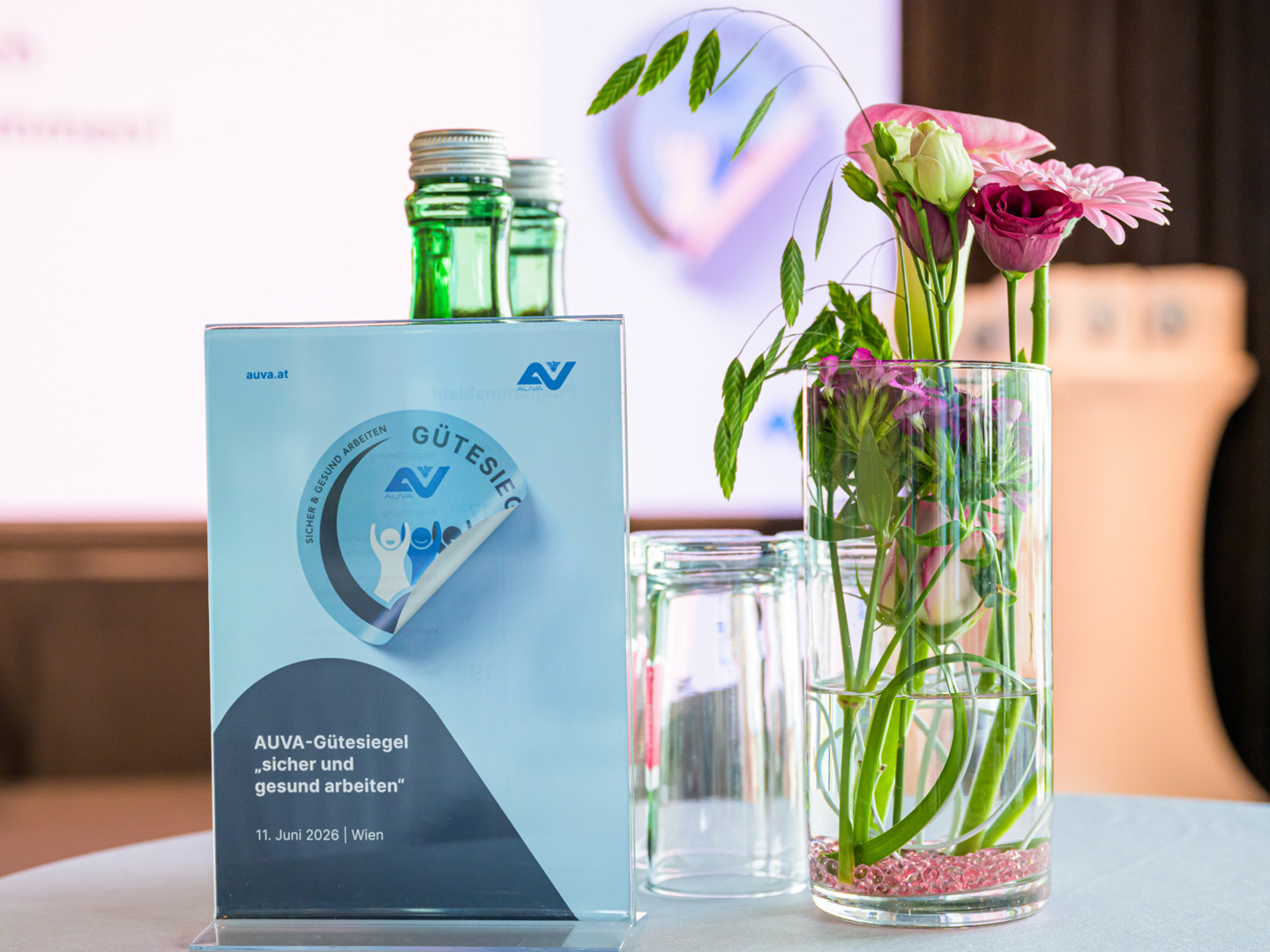 Nahaufnahme eines Tischaufstellers zur AUVA-Gütesiegel-Verleihung und Blumen in einer Vase