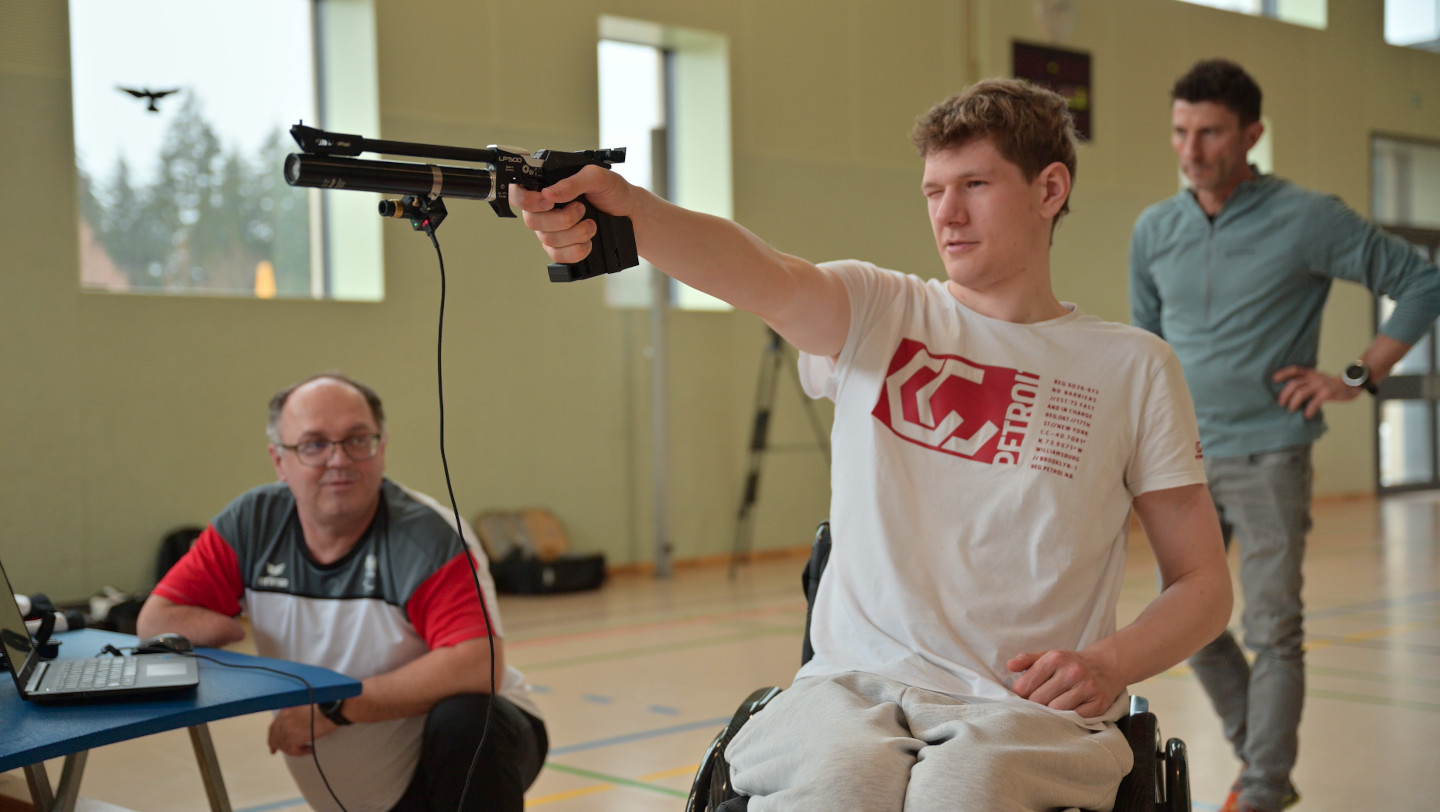 Mann im Rollstuhl zielt mit einer Laserpistole, links und rechts von ihm sieht man zwei Betreuende