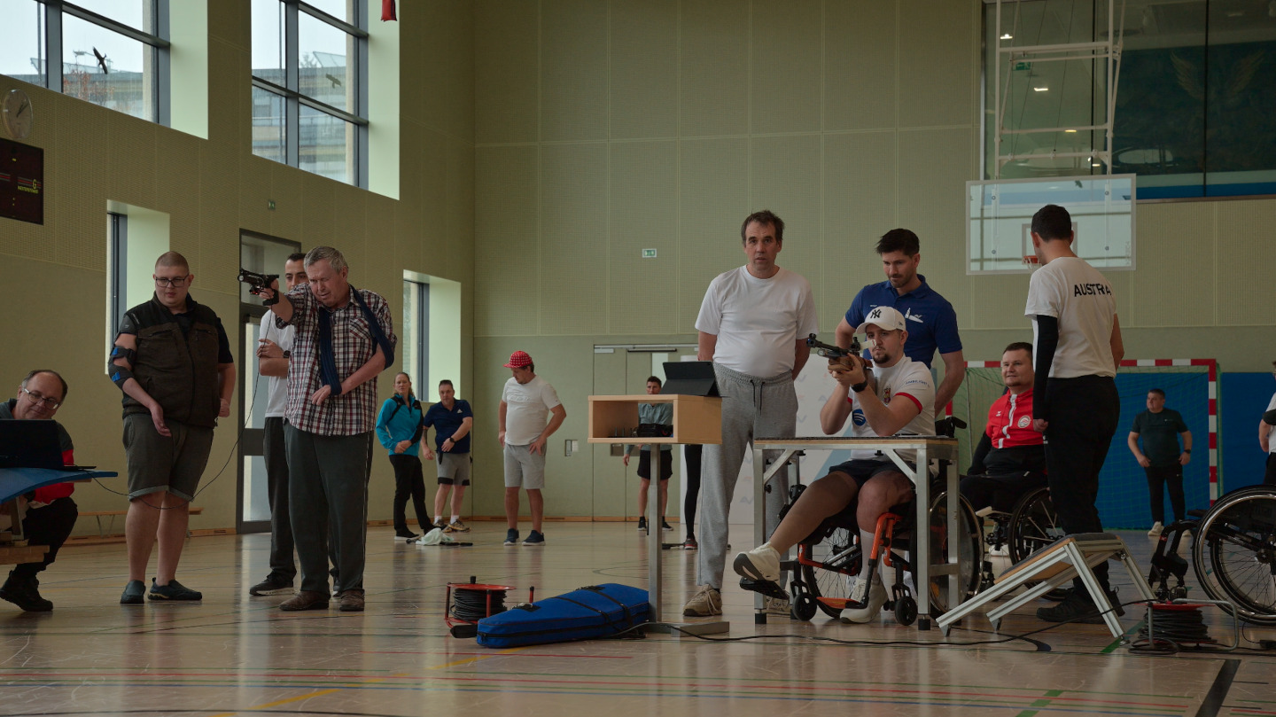 Zwei Männer zielen in einem Turnsaal mit Lasergewehren, um sie herum sieht man einige Betreuende