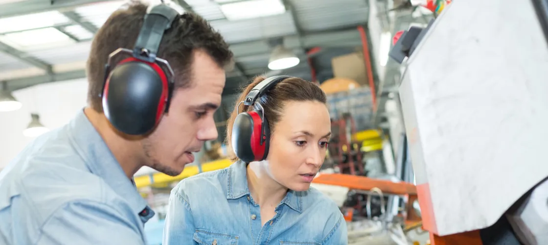 Adobestock 421177559 Gehoerschutz 1440 C Auremar S Ein Mann und eine Frau mit Kapselgehörschutz inspizieren eine Maschine in einer Produktionshalle.