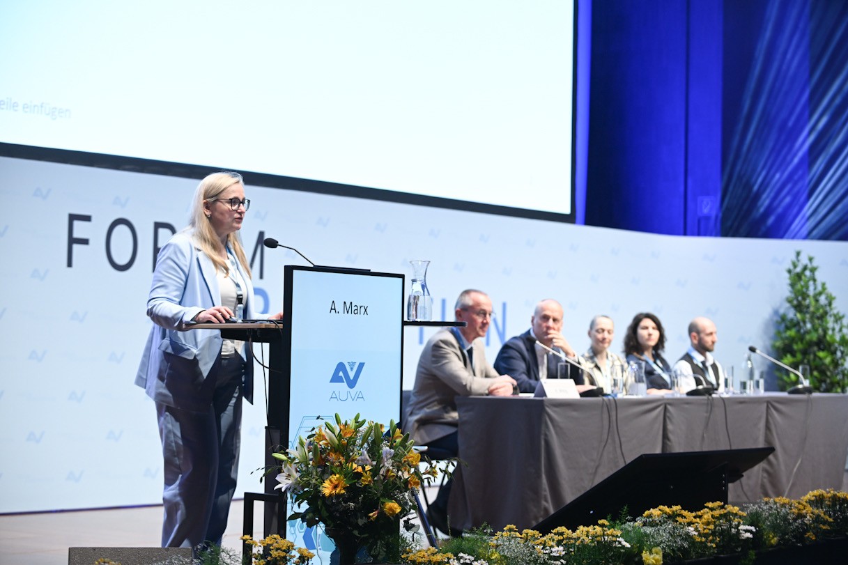 Eine Frau steht hinter einem Rednerpult, daneben sieht man ein Podium mit fünf Personen