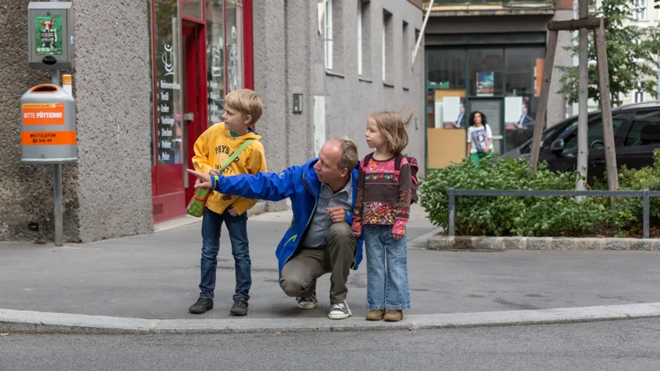 Schulweg 1440 Foto P.Winkler AUVA Mann hockt zwischen zwei Kindern am Gehsteigrand und deutet auf die Straße