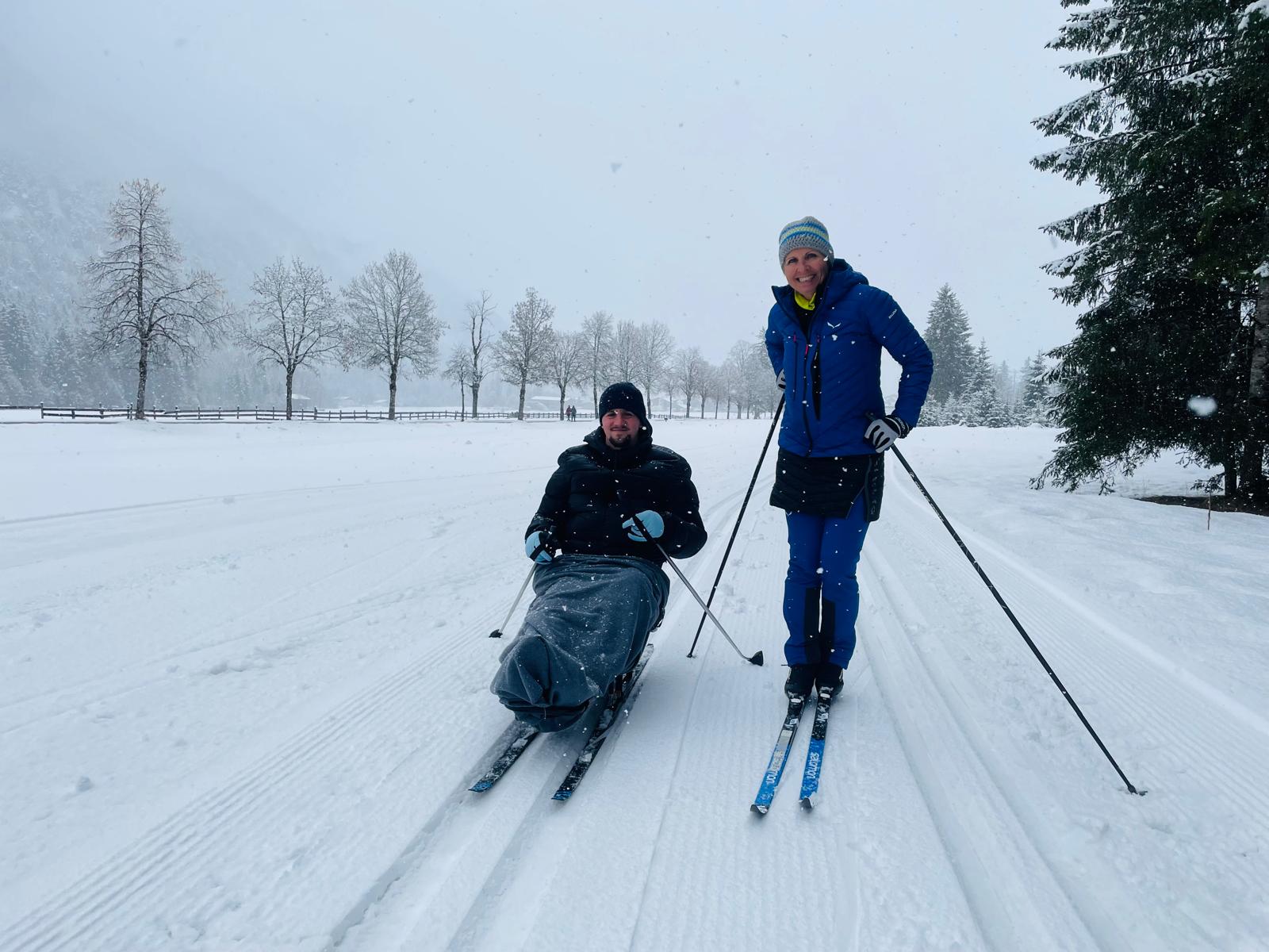 Links im Bild ein Teilnehmender auf einem Langlaufschlitten im Schnee, rechts im Bild ist die Trainerin zu sehen