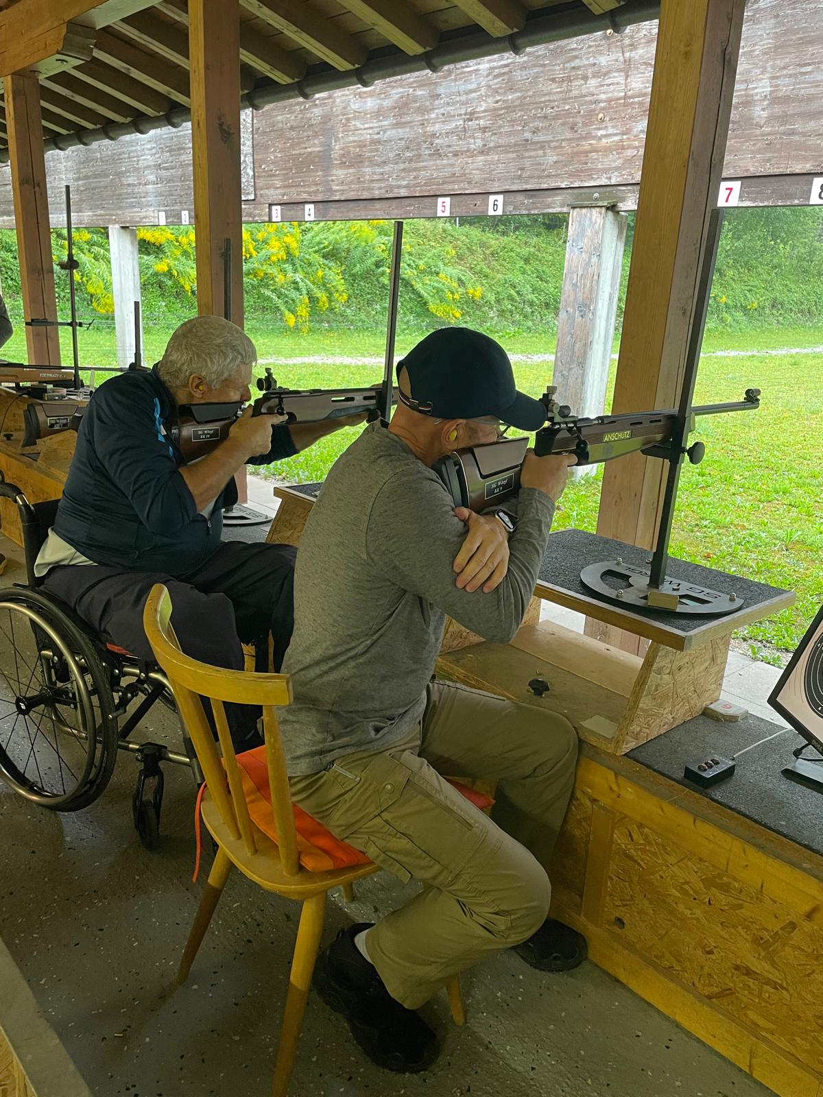 Zwei Teilnehmer sitzen im Schießstand und zielen mit dem Gewehr auf ein nicht erkennbares Ziel außerhalb des Schießstandes