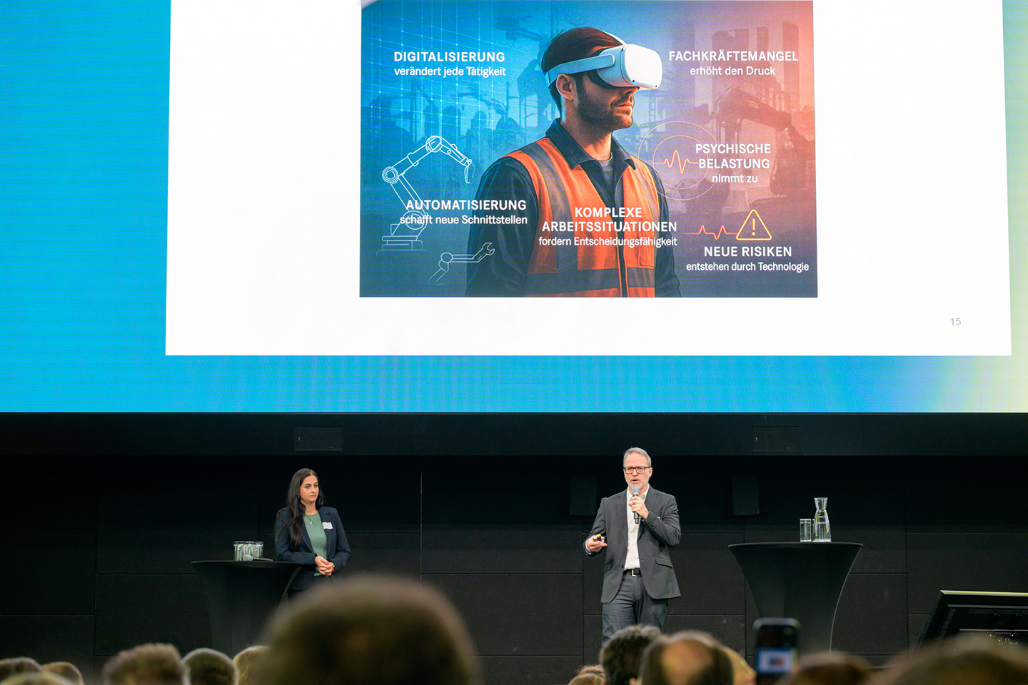 Ein Mann und eine Frau stehen auf der Bühne. Über ihnen ist eine Präsentation zu sehen mit einem Mann, der eine VR-Brille aufhat