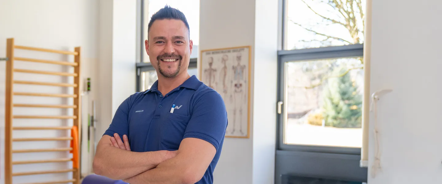 Ein Physiotherapeut steht mit verschränkten Armen vor zwei Liegen, im Hintergrund ist auch eine Sprossenwand