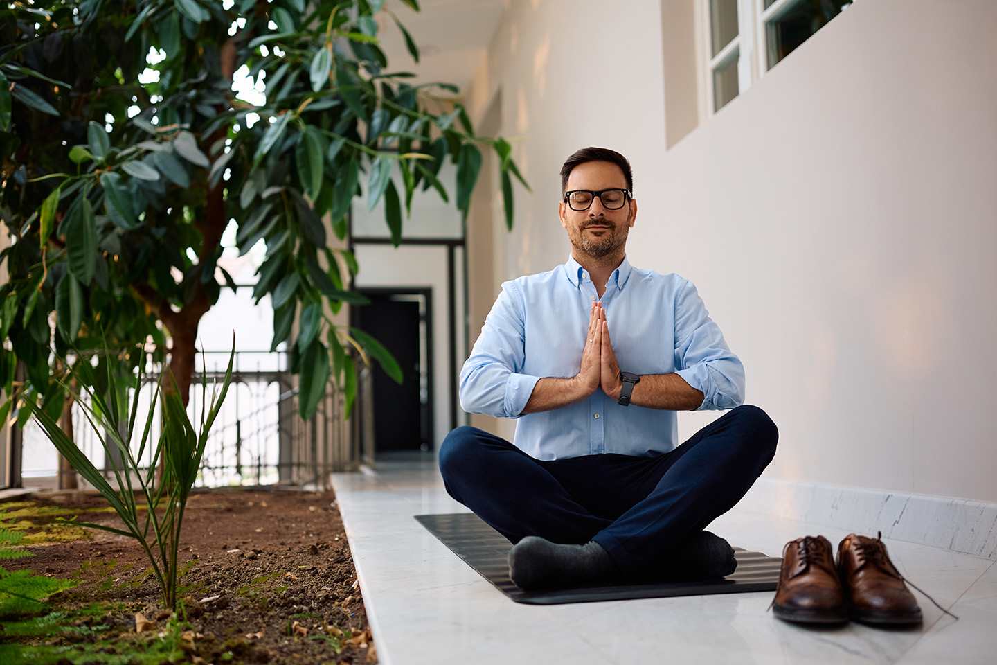 Ein Mann im Businessoutfit sitzt im Schneidersitz auf einer Yogamatte.