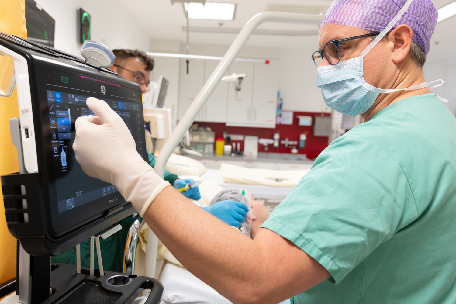 Mitarbeiter des Unfallkrankenhauses macht in einem Behandlungsraum eine Eingabe an einem Touchscreen, im Hintergrund ist ein weitere Mitarbeiter erkennbar