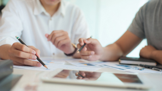 Zwei nur halb sichtbare Personen arbeiten an mehreren auf einem Schreibtisch liegenden Dokumenten