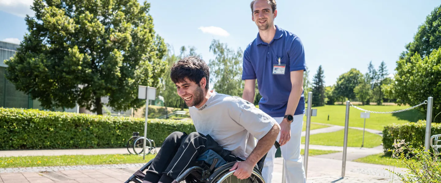 Patient mit einem Therapeuten beim Rollstuhltraining in den Parkanlagen des Rehazentrums Patient mit einem Therapeuten beim Rollstuhltraining in den Parkanlagen des Rehazentrums