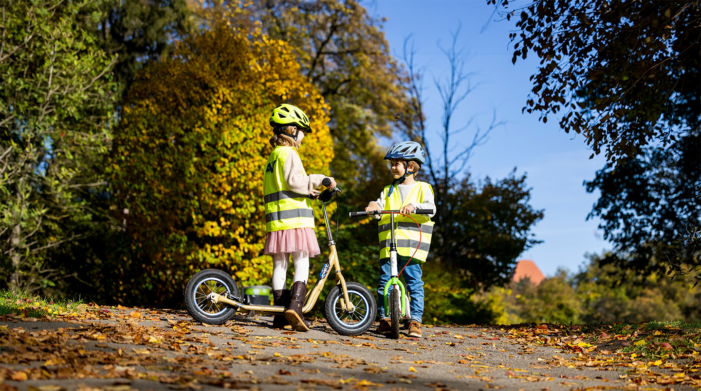 Ein Mädchen und ein Bub stehen mit Warnwesten und Helmen bekleidet mit ihren Tretrollern auf einem mit Herbstlaub bedeckten Weg.