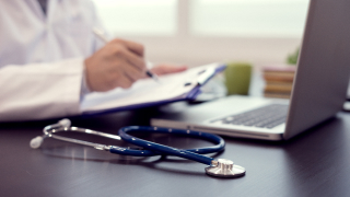 Ein Stethoskop liegt auf dem Schreibtisch vor einem nur halb sichtbaren Arzt, der am Laptop arbeitet