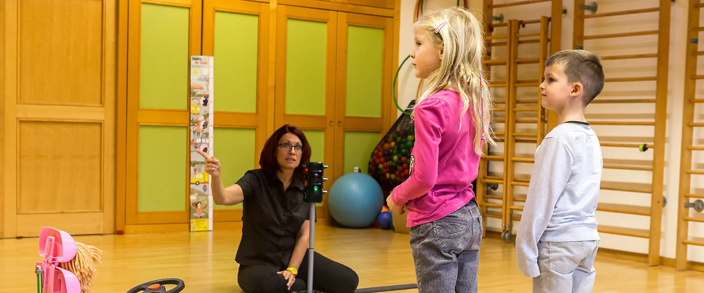 Zwei Kindern, einem Mädchen und einem Jungen, wird in einer Turnhalle von einer Lehrerin das richtige Verhalten in einer mit einem Bobby Car nachgestellten Verkehrssituation an einem Zebrastreifen erklärt.