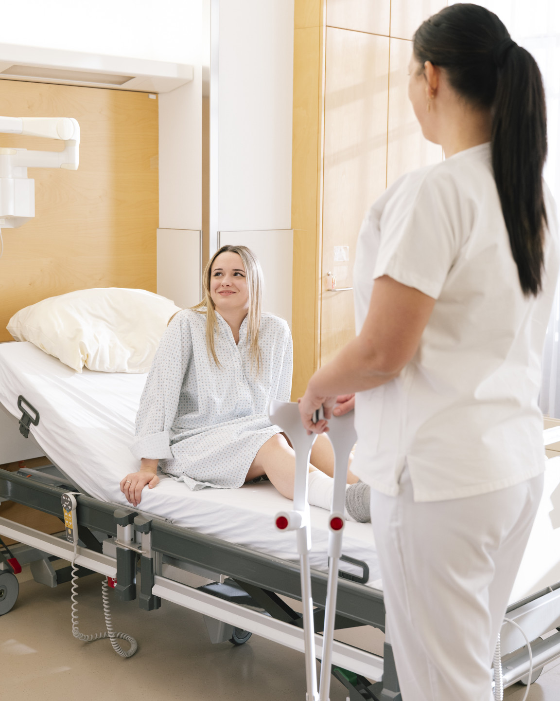 Eine Pflegerin steht mit Gehhilfen in der Hand vor dem Bett einer Patientin  