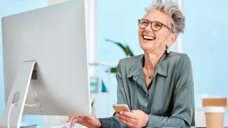 Fröhliche, erfahrene Frau am Bildschirmarbeitsplatz trägt eine Bildschirmarbeitsbrille.
