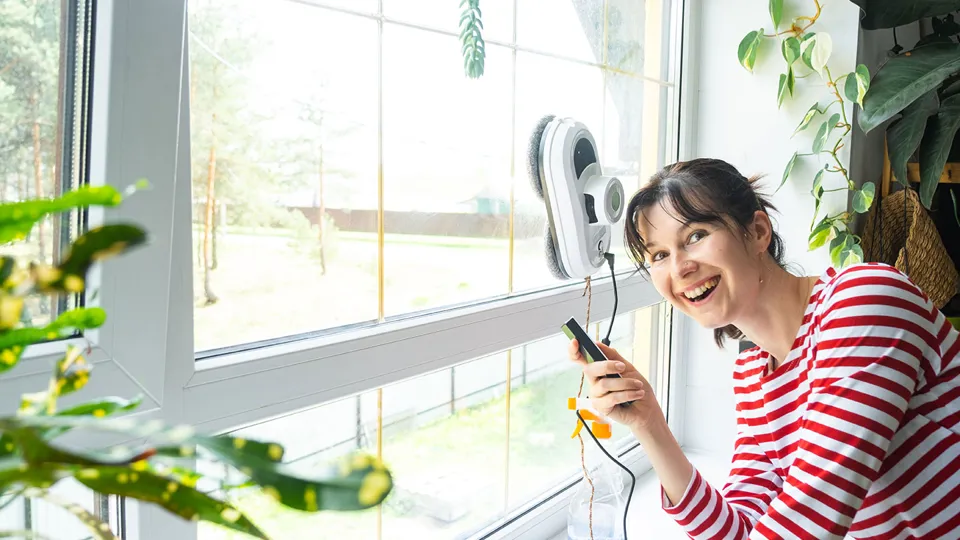 Eine lächelnde Frau bedient mit einer Fernbedienung einen Roboter zum Reinigen einer Fensterscheibe