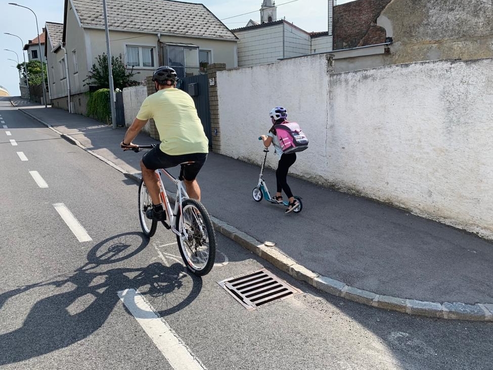 Ein Mann fährt auf einem Fahrrad auf einem Radweg. Daneben fährt ein Schulkind mit dem Tretroller am Gehsteig.