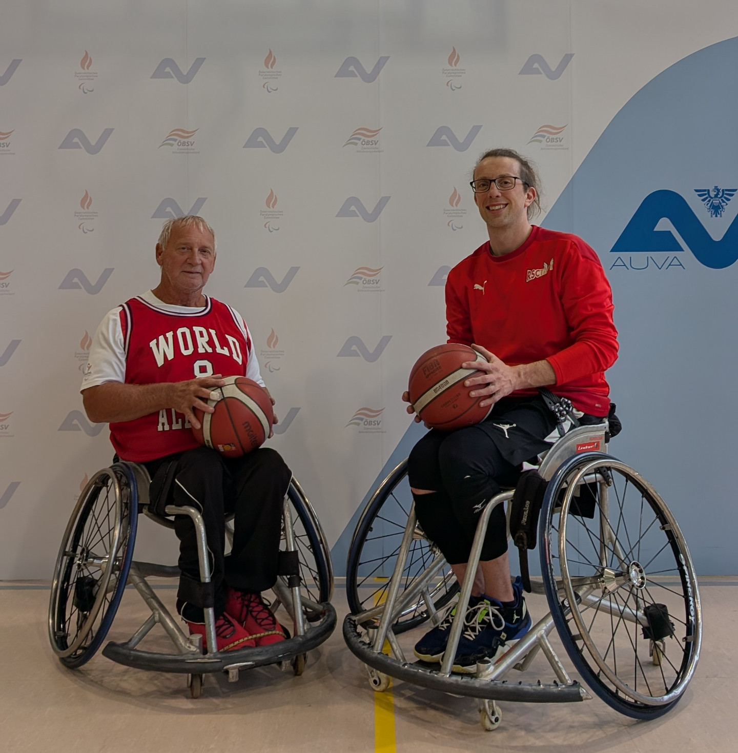 Zwei Rollstuhlbasketballspieler mit Bällen vor einer Wand mit AUVA- und ÖPC-Logos