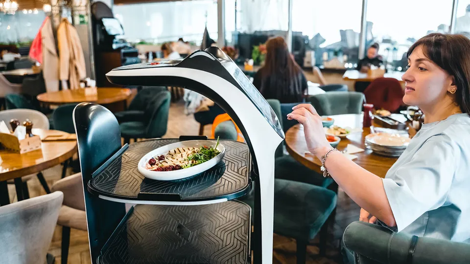 Eine Frau sitzt in einem Restaurant und schaut auf einen Servier-Roboter, der das Essen bringt.