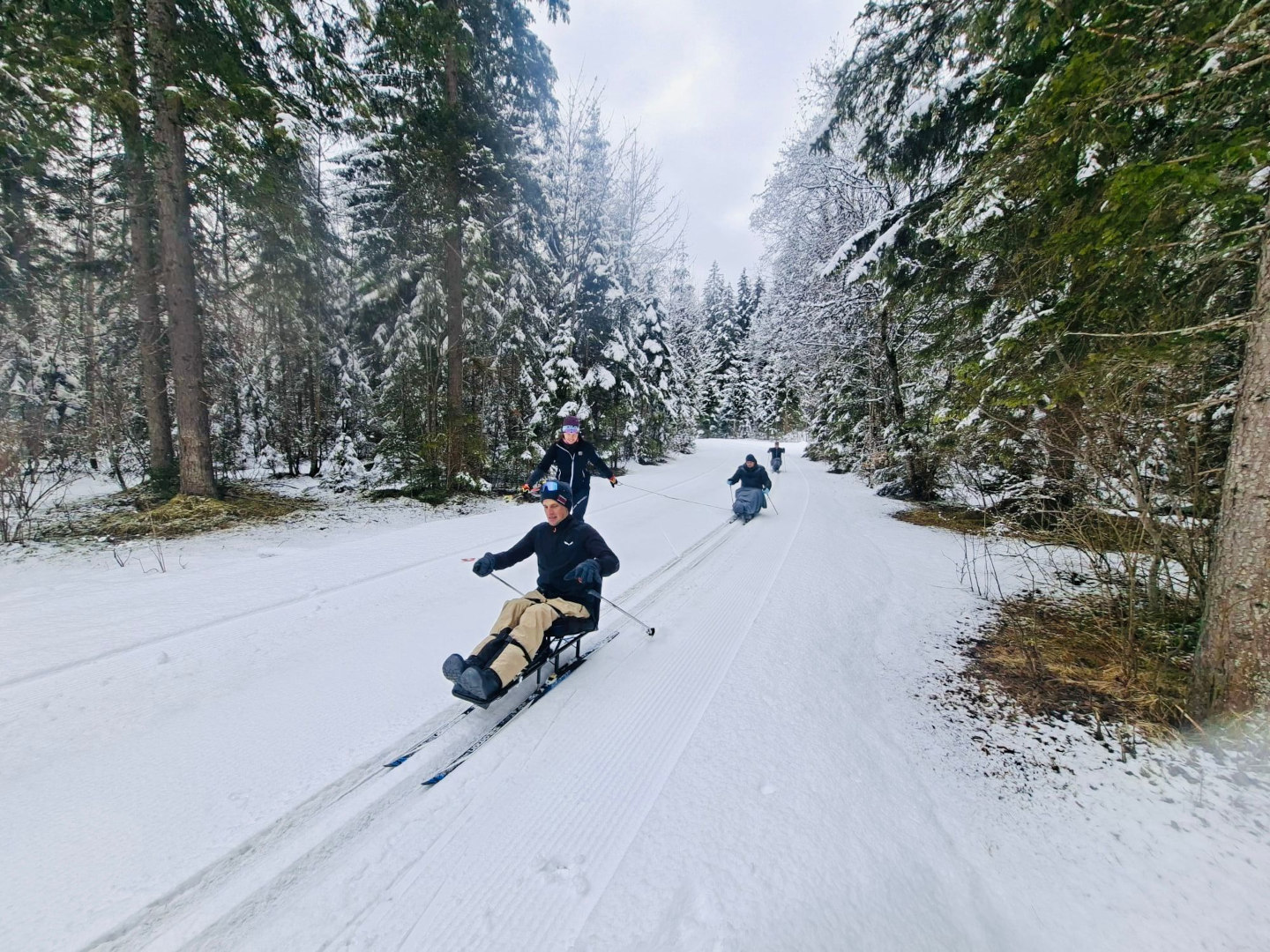 Drei Teilnehmende auf Langlaufschlitten im Schnee sind in Fahrt, im Bild ist auch die Trainerin zu sehen