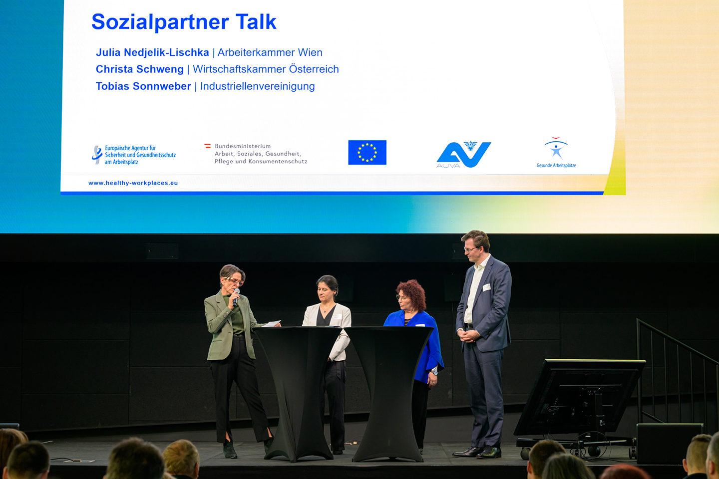 Vier Personen stehen auf einer Bühne. Eine spricht in ein Handmikrofon, die anderen hören zu.