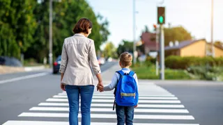 Schulweg C KI Adobe Firefly Edit Lwptm Rückansicht einer Frau und eines Kindes mit Schultasche. Sie stehen vor einem Zebrastreifen und blicken auf die gegenüberliegende grün leuchtende Fußgängerampel